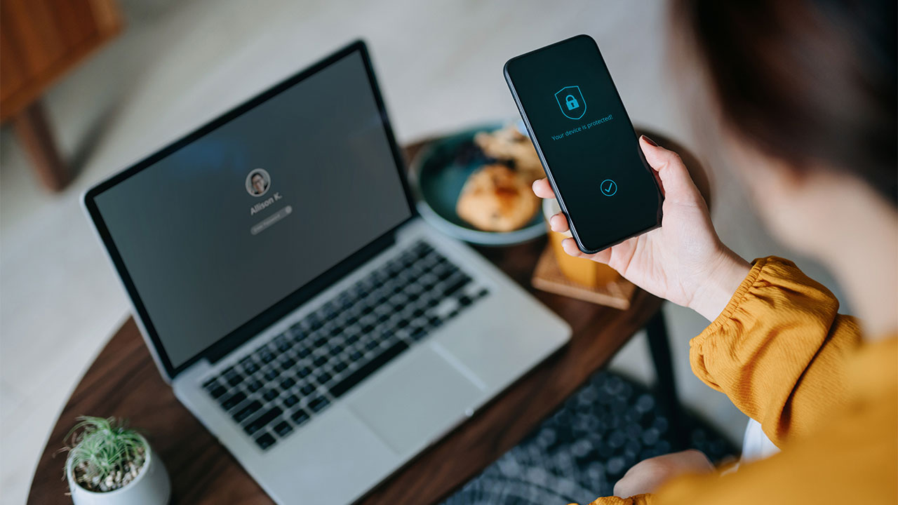 Person hält Smartphone mit Sicherheitsanzeige über einen Laptop mit Login-Fenster.